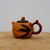 A detailed profile shot of a small, handcrafted Yixing clay teapot resting on a rustic, light-colored wooden surface. The teapot features a warm, ochre-colored body with a subtle horizontal ridge encircling its center. Its nature-inspired design includes a handle crafted to look like a gnarled, reddish-brown tree branch and a dark brown, textured lid with concentric circles. A small knob shaped like a peach, colored in both orange and reddish-brown, sits atop the lid. On the side of the vessel, three dark charcoal-colored leaves are intricately applied as if growing from the handle. The teapot’s short spout is elegantly integrated into the body with a small reddish-brown accent. This traditional piece of Chinese Zisha pottery is set against a clean, minimalist white background, highlighting the organic textures and fine craftsmanship of the clay.