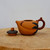 A high-resolution, side-view photograph of a small, artisanal Yixing clay teapot and its separate lid resting on a rustic, light-toned wooden surface. 

The main body of the teapot is crafted from a warm, golden-orange Zisha clay with a smooth, unglazed matte finish. Its organic shape is reminiscent of a plump peach or fruit, with a short, integrated spout. Decorative elements include dark, charcoal-colored clay leaves that wrap around the back near the handle and a small, reddish-brown bat or butterfly motif applied near the spout. 

The curved handle is made from a darker, reddish-brown clay, sculpted to look like a gnarled tree branch with a small knot on top. To the left, the matching lid is dark grey with a contrasting orange peach-shaped finial. The entire piece is set against a clean, neutral white background, emphasizing the intricate hand-carved details and traditional craftsmanship of this functional piece of Chinese pottery.