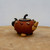 A small, artisan-crafted Yixing clay teapot with a nature-inspired design sits centered on a light-colored wooden surface. The teapot’s rounded body is a rich, warm reddish-brown with a smooth finish. It is uniquely decorated with several realistic, three-dimensional clay motifs that resemble various nuts and seeds, such as peanuts and beans, encircling its upper shoulder. 

The teapot features a short, stout spout made of a light tan, textured clay that mimics the appearance of a ginger root. Its handle is dark, almost black, and shaped like a gnarled, twisted vine. The flat-topped lid matches the body’s color and is crowned with a small, curved, twig-like handle in a dark finish. Instead of a standard base, the teapot is elevated by small, textured clay feet that look like individual nuts and seeds. The entire piece is captured against a plain, light-gray background, emphasizing the intricate, handmade details of the Yixing pottery.