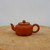A side-profile view of a traditional Yixing Zisha clay teapot, model YTP-04, showcasing its rich reddish-brown or terracotta hue. The teapot has a classic, minimalist design featuring a squat, rounded body, a short, elegantly curved spout, and a sturdy circular handle. Its flat lid fits snugly and is topped with a small, perfectly spherical knob. The unglazed clay surface has a smooth, matte finish that highlights the natural, fine-grained texture of the material. The teapot is centered on a rustic, light-colored wooden platform with visible grain and natural imperfections, set against a clean, neutral white background. This artisanal piece is ideal for traditional Gongfu tea service and serves as an elegant decorative accompaniment to a bonsai collection.