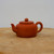 A profile view of a traditional handcrafted Yixing tea pot, model YTP-04, resting on a light-colored, rustic wooden slab. The teapot is made from fine, unglazed reddish-brown Zisha clay with a smooth, matte finish. It features a compact, squat, and rounded body shape with a short, elegant spout on the left and a thick, circular handle on the right. The flat-topped lid sits flush with the opening and is topped with a small, perfectly spherical knob. The background is a clean, minimalist off-white, highlighting the earthy tones and artisanal craftsmanship of this classic Chinese clay teapot.