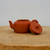 A small, handcrafted Yixing clay teapot in a rich reddish-brown terracotta color, featuring a smooth, unglazed matte finish. The teapot has a low-profile, rounded body with a curved upward-pointing spout on one side and a classic C-shaped handle on the other. Its matching circular lid is placed leaning against the right side of the pot, showcasing the small round knob on top which has a tiny steam vent hole in the center. The teapot sits on a thick, light-tan wooden pedestal that displays a natural wood grain texture. The entire scene is set against a simple, solid off-white background, highlighting the teapot's elegant and minimalist design as a functional piece of art or a decorative accessory for a bonsai display.