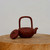 An elegant, handcrafted Yixing teapot made from traditional reddish-brown Zisha clay sits atop a rustic wooden pedestal. This specific piece, often used in tea ceremonies or as a bonsai companion, features a prominent overhead handle, known as the 'Ti Liang' style, which forms a tall, rounded rectangular arch over the pot's body. The side of the rounded teapot is delicately hand-etched with a subtle, traditional Chinese landscape scene of misty mountain peaks. Its matching circular lid rests on its side to the left of the pot, revealing a small knob with a steam vent. A short, gracefully curved spout extends from the right side. The entire arrangement is photographed against a clean, neutral white background, highlighting the earthy tones and fine craftsmanship of the ceramic art.