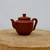 A detailed studio photograph of a traditional Yixing teapot (model YTP-06), elegantly showcased on a rustic, thick cross-section of a light-colored wood log. The teapot is crafted from authentic, reddish-brown Zisha clay, featuring a smooth, unglazed, and matte finish that highlights the natural beauty of the material. Its design is minimalist and well-balanced, consisting of a rounded, bulbous body, a short, gracefully curved spout, and a sturdy loop handle. The well-fitted, flat-topped lid is finished with a simple, cylindrical knob for easy handling. Set against a clean, off-white background with soft, even lighting, the earthy tones of the teapot and the natural wood base evoke a sense of calm and traditional craftsmanship, reflecting the deep connection between Yixing pottery, Chinese tea culture, and the aesthetic world of bonsai.