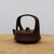 A side-profile view of a contemporary Yixing purple sand (Zisha) clay teapot with a matte, dark brown finish. This unique piece of teaware features a wide, low-profile body and a large, integrated overhead bridge handle that forms a bold, rounded arch over the center. A small, simple loop handle sits on the flat lid nestled beneath the main arch. The short spout curves gently upward from the side. The teapot is presented on a rustic, light-colored wooden surface against a clean, neutral white background, highlighting its smooth texture and minimalist aesthetic. This artisanal Chinese teapot blends traditional craftsmanship with a modern, architectural form, making it a functional piece of art for any tea ceremony.