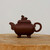 An image of a traditional Yixing clay teapot, a classic example of Chinese functional art, is shown from a side profile. The teapot is crafted from a deep, matte reddish-brown Zisha clay, prized for its ability to enhance the flavor of tea over time. 

The body of the teapot features a soft, rectangular form with rounded edges, elegantly supported by four delicate feet that curl slightly outwards at the base. A matching lid sits perfectly on the rim, topped with a finely sculpted decorative knob in the shape of a reclining mythical creature, such as a shishi or lion dog. A short, upward-curving spout is balanced on the opposite side by a smooth, rounded handle, designed for both aesthetic harmony and ease of use. 

Set upon a thick, natural wooden surface against a clean, off-white background, the teapot’s smooth yet earthy texture is highlighted by soft, even lighting. This piece exemplifies the craftsmanship of Yixing pottery, making it a sought-after item for tea enthusiasts and a beautiful accessory for bonsai displays or traditional tea ceremonies.