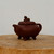 A handcrafted, dark reddish-brown Yixing clay teapot featuring a bulbous, four-sided body with softly rounded corners. The teapot stands on four short, elegantly curved legs, a design reminiscent of an ancient Chinese bronze ding. The flat, square-ish lid is topped with a finely detailed miniature sculpture of a guardian lion, or Pixiu, which serves as the decorative knob. A short, thick spout extends from one side, balanced by a simple, smooth C-shaped handle on the opposite side. The teapot's matte, unglazed surface highlights the natural, fine-grained texture of the traditional zisha clay. It is displayed on a light-colored, rustic wooden pedestal against a minimalist, neutral off-white background, emphasizing its classic form and artistic craftsmanship.