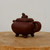 A detailed side profile of a traditional Chinese Yixing teapot made from dark reddish-brown Zisha clay. The teapot features a square-shaped body with rounded corners, supported by four small, dainty curved feet. Its square lid is topped with a finely detailed sculptural knob shaped like a small lion or mythical creature. A wide, rounded handle is positioned on the right side, balanced by a short, sturdy spout on the left. The teapot sits on a light-colored, circular wooden cross-section against a neutral, off-white background, highlighting the smooth, matte texture of the unglazed clay. This handcrafted ceramic piece is a staple of authentic tea culture and serves as both a functional tea vessel and a decorative art object.