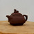 A detailed side-profile view of a traditional dark brown Yixing clay teapot (model YTP-13) resting on a light-colored natural wood surface against a plain white background. The teapot is crafted from unglazed, dark-toned zisha (purple sand) clay, giving it a matte, earthy texture characteristic of authentic Yixing pottery. Its unique architectural design features a squat, square-based body with a prominent horizontal ridge encircling the middle. A short, elegantly curved spout emerges from one side, while a sturdy, D-shaped handle is positioned opposite. The tiered lid mirrors the square shape of the body and is adorned with a meticulously carved finial shaped like a mythical Chinese creature, such as a Pixiu or a guardian lion, sitting in a watchful pose. This artisanal tea set piece exemplifies the classic, functional elegance of Yixing teapots, which are highly prized by tea enthusiasts and often used as decorative accents in bonsai displays.