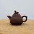 A side-profile shot of a handcrafted, square-shaped Yixing teapot made from rich, dark-brown Zisha clay. The teapot features a distinct geometric design with a robust, rectangular body and a matching tiered lid. Atop the lid sits a small, detailed figurine of a mythical creature, serving as a decorative finial. The teapot’s design is completed by a short, tapered spout and a thick, angular C-handle that follows the vessel's square theme. This functional piece of traditional Chinese art is displayed on a light-colored, rustic wooden cross-section against a clean, neutral white background, highlighting its earthy texture and precise craftsmanship.
