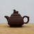 A profile view of a traditional square-shaped Yixing Zisha clay teapot in a deep, matte reddish-brown. The teapot rests on a light-colored, natural wood surface against a plain white background. Its design features horizontal decorative ridges around the top of the body, a short, upward-pointing spout, and a large circular handle with a decorative point on top. The matching square lid is multi-tiered and topped with a small, intricately carved finial in the shape of a mythical creature, possibly a lion or dragon. The unglazed purple clay texture is smooth yet earthy, characteristic of high-quality Yixing pottery used in traditional Chinese tea ceremonies.