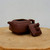 A side-angle, close-up photograph features a handcrafted square Yixing teapot (model YTP-13) made from traditional dark reddish-brown Zisha clay, also known as purple sand clay. The teapot has a geometric, boxy body with slightly curved walls, a short, upward-curving spout, and a sturdy, angular handle. Its matching square lid is propped against the right side of the teapot's base, showcasing a detailed sculptural ornament of a small mythical creature, like a qilin or lion, perched on top as the knob. A tiny steam hole is visible near the creature's base. The entire set rests on a rustic, light-colored wooden surface that looks like a cross-section of a tree trunk with visible natural rings and textures. The background is a minimalist, plain off-white wall, emphasizing the earthy tones and clean lines of the ceramic piece. This type of unglazed Yixing teapot is highly valued in Chinese tea culture for its ability to absorb tea oils and enhance flavors over time.