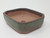 A high-angle, three-quarter view of a rectangular, cut-corner bonsai pot set against a plain white background. The pot features a matte, weathered green-finished exterior that blends with patches of unglazed reddish-brown clay, giving it an antique look. The top rim is highlighted in a dark chocolate brown, while the interior is the same natural terracotta-colored clay. In the bottom of the pot, a large central drainage hole and a smaller wire hole are visible. The pot stands on four small feet at the corners, which elevate it slightly for better drainage and air circulation. Its robust and earthy design makes it ideal for a variety of bonsai tree styles.