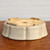 A detailed close-up shows a cream-colored glazed ceramic bonsai pot, positioned upside down to highlight its underside and construction. The pot features a classic Mokkou style with a soft, scalloped or lobed rim that creates a rhythmic, flower-like silhouette. 

The exterior is coated in a smooth, semi-matte off-white glaze that transitions into the unglazed, natural tan clay of the bottom. The underside reveals a large circular drainage hole at the center and four smaller holes designed for securing bonsai wire. Four low, sturdy integrated feet are spaced around the base to provide essential elevation for air circulation and drainage. The pot sits on a textured, reddish-brown wooden surface against a clean, neutral white background, emphasizing the craftsmanship of the pottery.
