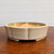 An eye-level studio photograph features a shallow, glazed ceramic bonsai pot sitting on a polished dark wood surface against a plain white background. The pot is finished in a soft cream or off-white glaze with a subtle matte texture. Its design features a scalloped, flower-like rim with vertical ridges along the sides, giving it a lotus or chrysanthemum shape. The interior of the pot shows a transition into a golden-tan or light amber glaze, adding a warm contrast to the exterior. The pot rests on low, integrated feet that provide a slight lift from the wooden table. The lighting is soft and even, highlighting the smooth curves and architectural details of the vessel, which is designed for housing small to medium-sized bonsai trees.