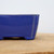 A detailed, close-up side view of a rectangular ceramic bonsai pot featuring a vibrant, high-gloss cobalt blue glaze. The image highlights the pot's smooth finish, rounded corners, and small, integrated 'cloud' feet. A slight reveal of the lighter-colored clay body is visible along the edges and corners, adding character to the piece. The pot, identified as model YX1113B from Bonsai Outlet, rests on a textured light brown wooden surface against a minimalist, out-of-focus white background.