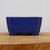 A front-view photograph of a small, rectangular ceramic bonsai pot with a vibrant cobalt blue glazed finish. This artisan bonsai pot, model YX1113B from Bonsai Outlet, features slightly flared sides and four small, integrated blocky feet at each corner that lift the base for drainage. The deep blue glaze has a high-gloss sheen with subtle color variations and minor wear along the top rim where the lighter clay body is slightly visible, giving it a handcrafted character. The pot is positioned in the center of the frame, resting on a light-toned natural wood surface with a visible grain pattern. The background is a clean, neutral off-white, which makes the rich blue color of the glazed pottery stand out. This piece represents a classic style of glazed bonsai pots used to complement the aesthetic of miniature trees.