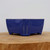 A small, deep blue glazed ceramic bonsai pot, model YX1113B, is shown from a side-on perspective. The pot has a rectangular form with slightly rounded corners and four short, blocky feet that elevate the base. Its surface is coated in a vibrant, glossy cobalt blue glaze that reflects the light, with thin lines where the lighter ceramic body shows through at the top rim and along the corners, adding a touch of rustic character. The pot sits centrally on a thick, textured light brown wood slice, set against a clean, off-white wall.