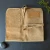 A top-down view shows a high-quality, tan suede tool roll by Tinyroots laid open on a dark, textured slate surface. The roll is made of a light brown suede with a soft, napped texture and rounded corners at the top.

A vertical row of small, silver metallic rivets runs down the center, while several horizontal pockets for holding tools are visible at the bottom. On the upper right side, a rectangular leather patch is stitched on, embossed with the "Tinyroots" brand name in a modern font. Two long suede straps are attached for tying the roll shut when it's folded up.

The scene is accented with natural elements: a small, bright green sprig of moss or fern is in the upper right corner, and a smooth, dark gray river stone sits near the bottom left corner, creating a rustic and professional aesthetic.