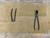 A high-angle, flat-lay photograph showcases a handmade tan suede leather roll-up tool bag from Tinyroots, designed specifically for organizing and protecting bonsai tools. The bag is spread wide open on a rustic, weathered wooden workbench. It features a series of vertical pockets of varying widths, each meticulously stitched and reinforced at the top opening with a small, circular silver metal rivet. 

Two professional-grade black metal bonsai tools are featured: on the left, the long, slender handles of a pair of concave cutters are tucked into one of the wider pockets, while on the right, a pair of black metal knob cutters with their distinctive rounded, biting jaws lies flat across the pocket area. The soft, textured surface of the tan leather provides a sharp visual contrast against the dark, matte finish of the precision maintenance tools. A large top flap made of the same light brown leather is visible above the pockets, intended to be folded down over the tool heads to keep them secure and prevent them from rubbing together when the bag is rolled up for transport or storage.