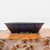 An elegant, shallow ceramic bonsai pot with a rich, mottled purple glaze is shown in a side-profile view. The glaze features a complex blend of deep violet, indigo, and dark charcoal tones, giving the handcrafted piece a textured, artisanal appearance. This oval-shaped pot has sides that flare gently outward toward the rim and is supported by four small, blocky feet at the base. It sits atop a thick, natural wooden slab with a prominent, knotty burl texture and warm golden-brown hues. The clean, neutral white background provides a sharp contrast, drawing focus to the unique colors and craftsmanship of this American-made Roy Minarai bonsai pot.