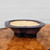 An exquisite, American-made Roy Minarai bonsai pot, showcased from a slightly elevated side angle. This handmade ceramic pot features a classic mokkou (four-lobed) oval shape with a wide, outward-flaring rim and sits on four sturdy, carved cloud feet. 

The exterior is coated in a striking, mottled glaze with deep shades of royal purple, dark burgundy, and indigo, creating a textured, organic look with crystalline-like patterns that catch the light. In contrast, the interior is unglazed, showing off the natural, pale tan color of the clay. The pot is artfully placed on a highly polished, warm-toned burl wood surface, with its rich colors reflected in the wood's grain, all set against a simple, off-white background with subtle horizontal lines. This piece represents the high-quality craftsmanship of American bonsai pottery.