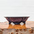 An American handmade ceramic bonsai pot, expertly crafted by artist Roy Minarai, sits centered on a polished wooden base. This shallow, mokkou-style (four-lobed) container features an elegant, outward-flaring rim and rests on four sturdy, integrated feet. The pot is finished with a striking, variegated glaze, showcasing a complex blend of deep indigo and shimmering violet tones with a mottled, almost crystalline texture reminiscent of traditional oil-spot techniques. It is displayed on a glossy, warm-toned burl wood slab with a rugged, natural bark edge in the foreground, all set against a clean, minimalist white background that emphasizes the rich colors and artisanal craftsmanship of the vessel.