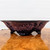 A profile view of a low, wide, handmade ceramic bonsai pot created by American artist Roy Minarai. The pot's exterior is adorned with a complex crystalline glaze featuring shades of lavender, deep purple, and rosy pink in a mottled, starburst-like pattern. Its flared rim tapers down to a base supported by three stout, rounded tripod feet finished in a dark, midnight blue glaze. The pot rests on a highly polished, honey-colored wood slab that shows a clear reflection of the vessel’s underside. The scene is set against a minimalist white background, emphasizing the intricate textures and artistic craftsmanship of this unique piece of bonsai pottery.