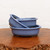 A professional product photograph showcases a nested set of three oval-shaped ceramic bonsai pots, ranging in size from 5 to 10 inches. The pots are finished in a smooth, glossy light blue or periwinkle glaze that catches the light along their rounded edges and distinct, slightly overhanging rims. 

The collection is artistically arranged on a warm-toned, polished wooden slab featuring a rustic, natural live edge at the bottom of the frame. The largest oval pot sits flat on the wood, serving as the base for the two smaller pots tilted inside it, highlighting their uniform color and shallow depth. The entire arrangement is set against a clean, minimalist white background, which emphasizes the cool blue tones and elegant silhouette of the pottery. Small, integrated feet are visible at the base of the pots, a functional design element essential for providing the drainage and aeration necessary for healthy bonsai tree growth.