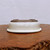 An upside-down, cream-colored glazed bonsai pot sits centered on a polished, warm-toned wood slab with a rustic, live edge. The pot features a smooth, rounded rectangular shape and is coated in a glossy, off-white glaze that has a subtle, brushed texture. Because it is inverted, the dark brown, unglazed clay base is clearly visible, showing four integrated corner feet and two large circular drainage holes essential for plant health. The simple, elegant design is set against a plain, light-colored background, emphasizing the pot's clean lines and neutral color palette, which would complement various bonsai tree species.