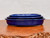 A side-profile view of three nested cobalt blue glazed ceramic bonsai pots, ranging in size from 10 to 15 inches, stacked on a polished, warm-toned live-edge wooden surface. The pots are oval-shaped with a shallow profile, characteristic of traditional bonsai containers, and feature a vibrant, high-gloss glaze that reflects light. The largest pot sits on the bottom, with two smaller but identically shaped pots tucked inside. The side view highlights the slightly flared rims and the sturdy, unglazed pale-gray base of the largest pot, designed for stability and drainage. The arrangement is set against a clean, minimalist off-white background, emphasizing the rich color and craftsmanship of the pottery.