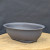 An eye-level shot captures a dark charcoal grey oval plastic bonsai training pot, which is centered on a light-colored circular wooden platform. The planter features a smooth, rounded form with a slightly flared upper rim and small, sturdy feet at the base for elevation and drainage. Its matte, textured surface gives it a professional appearance suitable for early-stage bonsai tree development. The pot is set against a dark, softly-lit grey fabric background, highlighting its simple and functional design. This type of durable plastic planter is a practical choice for training and growing bonsai trees before they are ready for a final decorative ceramic pot.