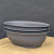 A set of three dark gray, oval-shaped plastic bonsai training pots stacked together on a natural wood surface. These durable, matte-finished planters are designed specifically for bonsai development and cultivation. Each shallow pot features small built-in feet for proper drainage and air circulation. The minimalist arrangement is set against a dark, textured background, emphasizing the functional design of these essential tools for bonsai enthusiasts and practitioners.