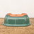 An upside-down rectangular bonsai pot rests on a rustic, weathered wooden tabletop against a clean, off-white background. The pot is finished in a vibrant, glossy seafoam green glaze that covers its decorative, lobed exterior walls and flared rim. Because it is inverted, the unglazed, reddish-orange terracotta base is clearly visible, showcasing functional features essential for bonsai care: a large circular central drainage hole and several smaller holes for threading bonsai wire to secure a tree. Four subtle, integrated feet are positioned at the corners of the base to provide elevation for airflow. The lighting creates soft reflections on the smooth, green glazed surface, contrasting with the matte, earthy texture of the ceramic bottom.