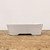 A studio photograph shows a side profile of a minimalist, rectangular glazed bonsai pot resting on a rustic, textured wooden tabletop. The ceramic pot features a smooth, matte white glaze with subtle variations, giving it an elegant and clean appearance. Its design includes slightly tapered walls that widen toward the rim and two small, arched cutouts at the base that act as integrated feet for improved drainage and airflow. The pot is centered against a plain, off-white background, highlighting its simple lines and functional form, typical of the 5 to 10-inch glazed bonsai pots found at Bonsai Outlet.