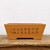 An eye-level, front-view photograph of a rectangular, unglazed ceramic bonsai pot made from warm, ochre-colored clay. The pot features a traditional tapered shape, narrowing slightly from the top rim down to a sturdy base with integrated feet visible as cutouts. The centerpiece of the design is a series of hand-etched Chinese calligraphy characters across the front face, rendered in a dark, contrasting tone that highlights the fine carving. The pot sits centrally on a rustic, weathered wooden surface with a prominent grain, all positioned against a stark, plain white background that emphasizes the earthy tones and textures of the unglazed stoneware.