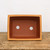 An overhead view looks into a rectangular, unglazed terracotta bonsai pot, emphasizing its deep interior and essential drainage features. The pot is crafted from reddish-brown clay with a contrasting lighter, sandy-tan rim that curves slightly outward at the top. At the center of the smooth, dark-brown interior base are two large, perfectly circular drainage holes, which are crucial for maintaining healthy bonsai root systems by preventing waterlogged soil. The pot sits on a rustic, weathered light-brown wooden tabletop, showcasing the natural textures of both the unglazed ceramic and the aged wood. The entire scene is set against a clean, minimalist white background, highlighting the simple, functional design of this professional-grade unglazed bonsai container.