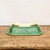 An upside-down view of a rectangular ceramic bonsai pot, showcasing its detailed construction and vibrant emerald green glaze. The pot features a beautiful crackle finish (crazing) across its exterior, which adds depth and an aged character to the piece. It is supported by four ornate, scroll-carved feet, also finished in the same rich green glaze. The flat, unglazed bottom reveals the natural light beige clay and includes several drainage and wire holes essential for proper bonsai plant care. The pot is displayed on a rustic, weathered wooden surface against a plain, off-white background, highlighting its elegant shape and classic bonsai aesthetic.