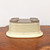 A close-up, high-angle shot shows an upside-down cream-colored glazed ceramic bonsai pot resting on a polished, dark burl wood surface against a stark white background. 

The pot is oval-shaped with a soft ivory or pale cream glaze that features a subtle, hammered texture along its sides, giving it a handcrafted appearance. Because the pot is inverted, the wide, flared rim is at the bottom, while the unglazed base faces upward. 

The base of the pot reveals the natural, earthy-gray clay body. It is designed for functionality, featuring two large circular drainage holes and four integrated, raised feet that allow for proper water runoff and aeration when in use. The neutral cream glaze and classic oval silhouette make this container a versatile choice for a wide range of bonsai tree species.