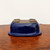 An upside-down view of a deep cobalt blue, rectangular glazed bonsai pot with rounded corners, resting on a polished reddish-brown wood surface against a plain white background. The image highlights the pot's glossy, reflective finish and structural details. The bottom is unglazed, showing the natural light-brown ceramic material, and features four integrated feet at the corners and large drainage holes essential for bonsai health. The prominent flared rim and elegant curves of the pot are clearly visible from this angle, showcasing its craftsmanship and design.