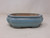 An eye-level, side-view photograph of a 7-inch glazed Yixing bonsai pot in a soft, light blue color. This shallow ceramic planter features a traditional mokkoh or scalloped rectangular shape with four rounded, lobed corners. The glossy cerulean glaze has a smooth finish with subtle dark speckles and a slightly weathered texture. 

The pot's design includes a wide, flat rim and a curved body that tapers slightly toward the base, resting on small, integrated feet. The unglazed interior reveals a natural buff-colored clay, providing a porous surface suitable for bonsai root health and drainage. The pot is presented against a plain, neutral white background, highlighting its elegant shape and serene blue hue.