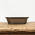An eye-level, full-shot photograph features a rectangular unglazed bonsai pot crafted from dark brown ceramic with a smooth, matte finish. The pot is characterized by its traditional design, featuring a flared rim, an elegant recessed rectangular panel on the side, and cut corners that create an octagonal silhouette. It rests on a tiered base with integrated small feet, designed to provide stability and proper drainage for a bonsai tree. The container is centered on a rustic, weathered wooden surface, which contrasts with the pot's clean lines. The entire scene is set against a stark, minimalist white background that highlights the rich, earthy tones and fine craftsmanship of this unglazed bonsai container.