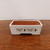 A rectangular white glazed bonsai pot with a floral motif sits on a polished dark wood surface against a plain light background. The planter features a clean, white exterior glaze that contrasts with the earthy, unglazed terracotta-colored interior. 

The front side of the pot is decorated with a detailed floral pattern, including two large dark-centered flowers that resemble sunflowers or dahlias, interspersed with clusters of smaller white and yellow blossoms and delicate green leaves. Beneath the flower clusters, the name "Frida Kahlo" is printed twice in a black stylized script font. 

The pot has slightly tapered sides and a flat rim, and it rests on two integrated feet created by a large rectangular cutout at the base to allow for drainage. The overall aesthetic combines a classic bonsai pot shape with modern, artistic floral elements.