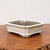 A rectangular white glazed bonsai pot with indented corners sits on a rustic, warm-toned wooden slab. The pot features a clean, minimalist design with a creamy, off-white glaze that has subtle, fine dark speckles, giving it a handcrafted ceramic appearance. 

The container has a wide, flat rim and matching raised detailing along the base, resting on four low-profile, cloud-style feet at the corners. While the exterior is smooth and glossy, the interior is unglazed, showing the natural dark brown clay body, which is ideal for root health and drainage. The pot is centered against a neutral, off-white background, highlighting its elegant shape and bright finish, which is traditionally used to provide a striking contrast for flowering or deciduous bonsai trees.