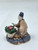 A small, handcrafted Chinese mudman figurine titled "Woman with Flowering Pot" (model F-044) sits against a plain white background. The clay figurine depicts a woman in a kneeling position, her body slightly angled. She wears simple, light brown traditional robes with dark blue-grey trim along the sleeves and neckline. Her head, made of white-painted clay, is tilted upwards, and she wears a large, dark-colored headpiece or bun adorned with a tiny red flower.

In front of her sits a small, white-ribbed pot overflowing with textured green foliage and several small pink-red blossoms. Her hands are raised slightly as if she is admiring or tending to the plant. The entire scene is mounted on a circular, dark blue-grey base with a ridged, segmented pattern. The figurine has the characteristic matte, earthy texture of traditional mudman art, often used as decorative accents for bonsai trees and miniature gardens.