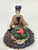 A detailed, handcrafted Chinese mudman figurine depicting a woman sitting behind a flowering pot. The miniature figure features a pale, serene face with simple painted features and dark hair styled in a traditional topknot, accented by a small red ornament. She is dressed in a tan or light brown robe with wide, dark-trimmed sleeves that have a subtle texture. Her hands are positioned as if she is carefully gesturing toward or tending to the plant in front of her.

The plant is held in a small, white, cross-hatched textured pot and consists of thick green foliage topped with vibrant pink or coral-colored flowers. The entire figurine is mounted on a circular, dark-colored base with a rough, stone-like texture and brown highlights. This rustic clay ornament, with its hand-molded characteristics, is a classic example of a Chinese mudman, often used as a decorative accent for bonsai tree displays or miniature indoor gardens to add a sense of life and tradition.