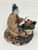 A detailed, handmade Chinese mudman figurine of a woman kneeling beside a flowering pot, set against a plain white background. The miniature figure is crafted in a traditional Shiwan style, featuring a blend of glazed and unglazed ceramic textures. The woman wears a textured, tan-colored robe with wide sleeves and black trim at the collar and cuffs. Her face is made of smooth white clay with simple, hand-indented features, and her black hair is styled in an ornate high bun adorned with a small red flower. She is positioned as if carefully tending to a white, fluted flower pot that contains a dense cluster of textured green foliage and bright pink blossoms. The entire scene sits upon a circular, domed base with deep, dark brown ridges that resemble a tortoise shell or rocky ground. This decorative figurine is a classic accessory for bonsai arrangements or miniature gardens.