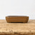 A front-facing, eye-level shot captures a rectangular, glazed bonsai pot in a warm, earthy brown tone. The ceramic pot features gently rounded corners and subtly fluted vertical lines at its edges, creating a soft, elegant silhouette. Its surface is finished with a smooth, slightly mottled glaze that catches the light, giving it a subtle sheen. The pot is elevated by four small, integrated feet at its base, designed to provide essential drainage and airflow for a bonsai tree. It sits centered on a rustic, light-colored wooden surface with visible grain and wear, set against a stark, plain white background that highlights the pot's rich color and clean craftsmanship.
