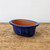 A deep cobalt blue glazed ceramic bonsai pot with an oval shape sits centered on a rustic, light-brown wooden surface. The pot features a rich, glossy blue glaze on its exterior and flared rim, with subtle dark mottling and vertical drip-like patterns adding texture to the finish. The interior of the pot is unglazed, revealing the natural, earthy reddish-brown terracotta clay. Small, integrated feet at the base elevate the container, providing space for drainage essential for bonsai tree health. The pot is positioned against a plain, off-white background that emphasizes its vibrant color and classic design, making it a professional-grade choice for showcasing a bonsai.