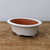 A shallow, oval-shaped white glazed bonsai pot sits centered on a rustic, weathered wooden table. The pot features a wide, smooth rim that curves outward and a contrasting unglazed reddish-brown terracotta interior. It is elevated slightly by four small, rounded white feet at the base. The exterior has a subtle, creamy matte finish with tiny, dark speckles. The background consists of a plain, neutral-toned wall, highlighting the clean lines and elegant design of this ceramic bonsai container.